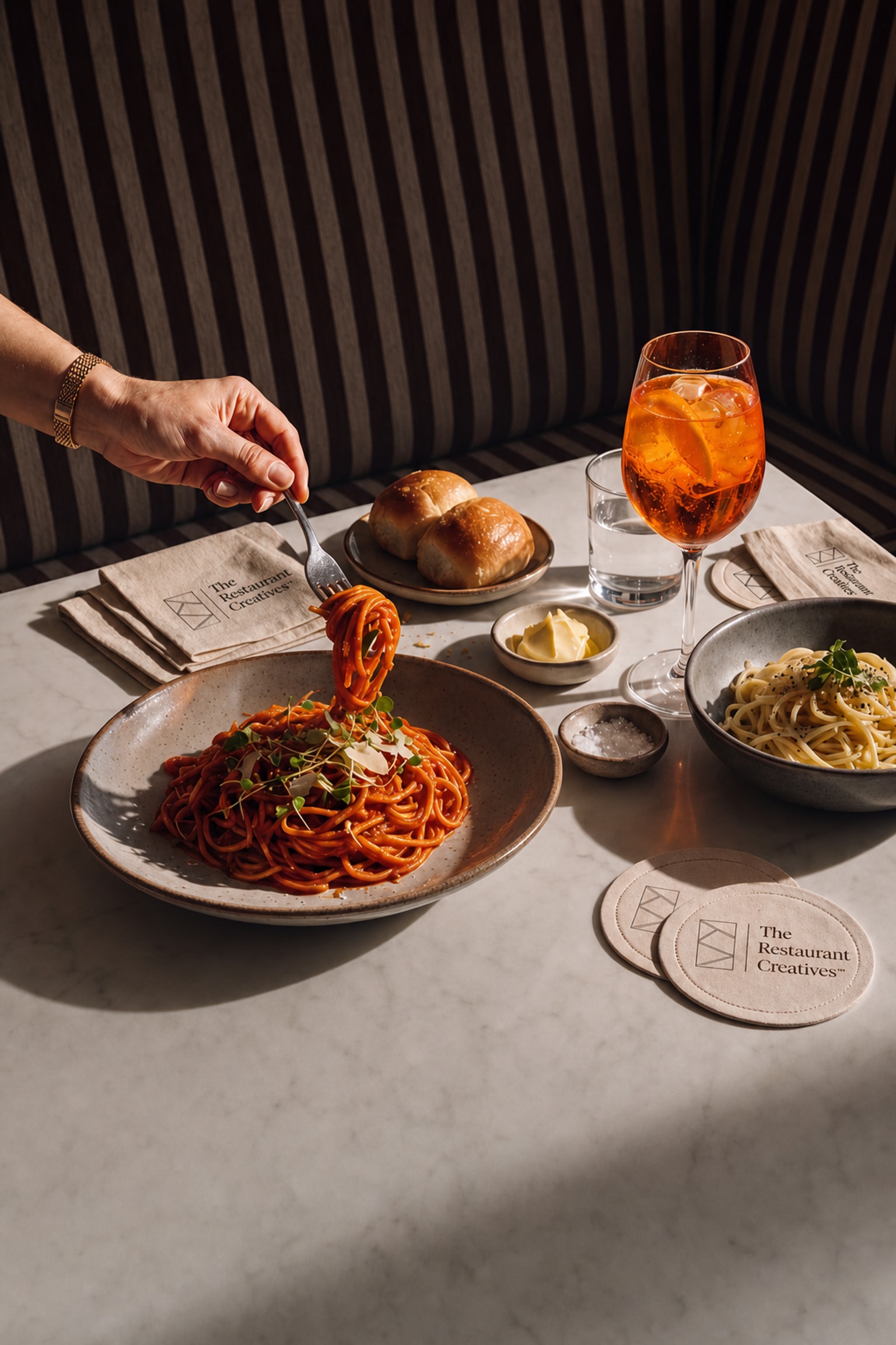 Restaurant table with pasta, Aperol spritz, bread rolls, and branded coasters at a striped booth